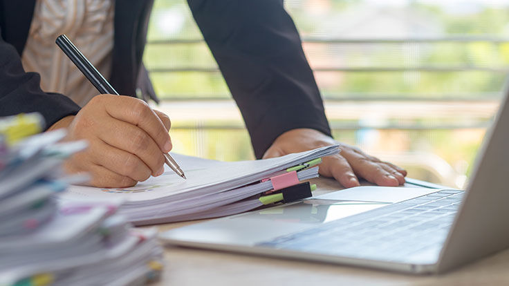 a person writing on paper with a stack of files on the table