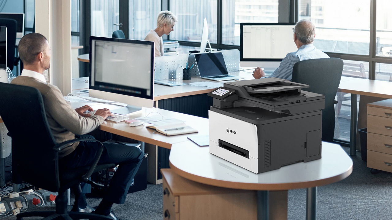 A Xerox C305 MFP printer on a desk in an office with 3 people